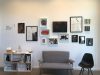 Art exhibition hung salon style on a white wall, a bookshelf and two chairs below the group of framed pieces.
