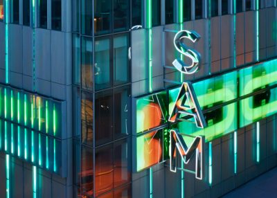 Corner of a modern, metal and glass building, with a neon sign reading "SAM"