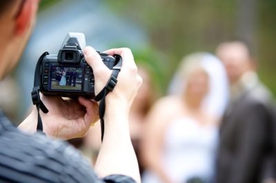 Close up of a DSLR camera screen over a photographer's shoulder, which shows a photo of a wedding scene.