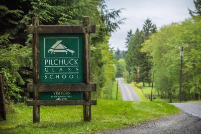 Wooden sign reading "Pilchuck Glass School", a two lane road in the background winding between pacific northwest forest.