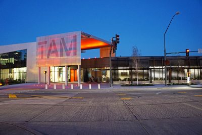 Exterior of a low glass and concrete museum, a large metal sign reading "TAM" over the entrance.