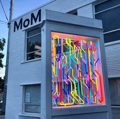 Neon light display in front of a white bricked building. Painted text on the outside of the building reads "MoM"