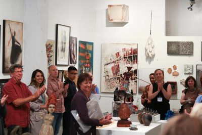 Group of people applauding a presentation in a gallery setting; work hung on the walls salon style and sculptural pieces displayed on pedestals.