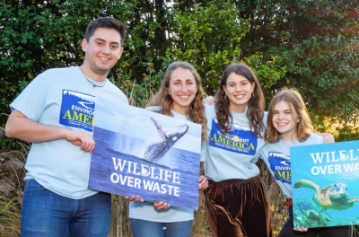 Group of 4 smiling youths of varying ages stand with arms over each others shoulders. They hold two signs with marine wildlife depicted and text reading "Wildlife Over Waste"