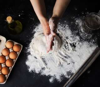 Aerial view of light skinned hands making pasta dough on a floured counter, a carton of eggs nearby.