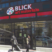 Exterior of a store entrance, sign above reading "Blick Art Materials." A bronze sculpture of a person playing guitar is in front of the entrance.