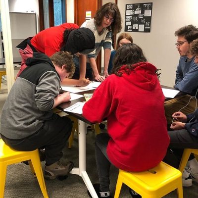 Group of teen youths work on drawings, gathered around a table in a classroom. An adult teacher leans over, listening to one of the students.