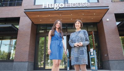 Two smiling femme people stand in front of a red bricked building entrance, a sign above reading "Hugo House"