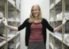 individual stands between two sets of metal shelving holding many museum art and anthropological objects. Individual is smiling, and wears curatorial gloves.
