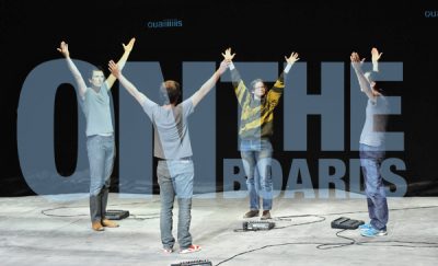 four people stand in a square formation on a stage, their arms raised. Translucent text imposed over the image reads ON THE BOARDS