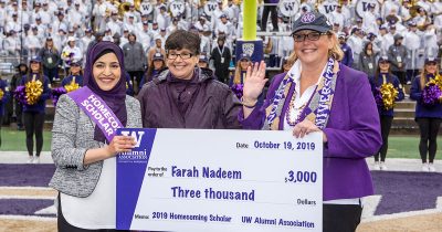 Three people stand on the field in a crowded sports stadium holding an oversized cheque; the person on the left wears a sash that reads "Homecoming Scholar"