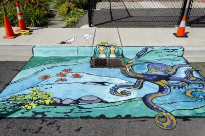 Painted ocean mural on a sidewalk and street, around a storm drain.