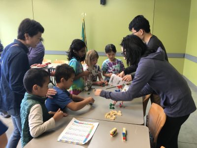 Young children gather around a table for a Lego-based activity, their parents standing around them.