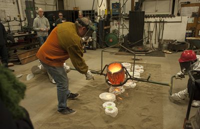 Two artists in a metalworking studio pour molten bronze into casts, while a group of people watch.