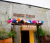 exterior of a grey concrete building, a sign reading "Might Tieton" on the front facade. A metal awning with multi-colored balloons over the entrance door. An orange banner hands from the metal awning readnig "10 x Tieton; Art Exhibition."