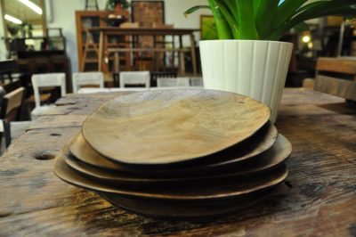 Hand carved wood plates rest on a wooden table, a plant in the background and other furniture placed in the space.