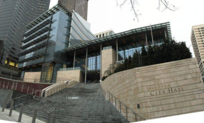 Exterior view of a modern glass building. Brick wall next to steps leading up to entrance with sign that reads "Seattle City Hall"