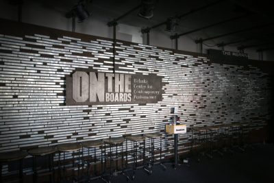 Interior accent wall with metal tile installation, a sign reading "On The Boards" in the center. Stools are lined in front of the wall.