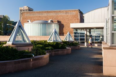 exterior view of a red bricked and metal museum. Three triangular metal sculptures line the walkway to the entrance. A person walks towards the front door.