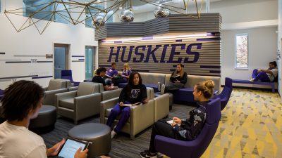 Students congregate in a lounge area full of purple and gold chairs and couches, a wall with the word 'Huskies' in purple lettering.