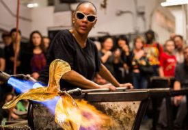 Woman with brown skin and sunglasses blows glass in front of an audience.
