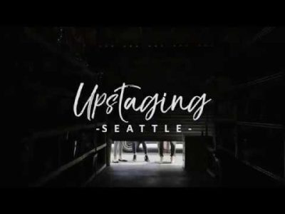 Darkened interior of a moving truck, the door just starting to open and let in light. Three sets of feet are visible on the outside. Text above door reads "Upstaging Seattle"