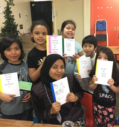 Group of school kids in varying ages, skin tones, and gender identities stand in a classroom setting. All of them hold small handmade books that have the same title - "My Hero"
