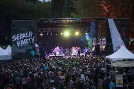 Large audience stands in front of a stage at an outdoor event venue. A band performs on stage, and a large banner to the left reads "Search Party"