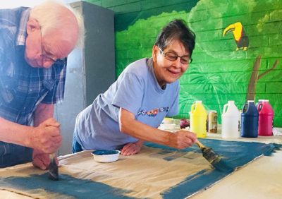 Two adults paint a piece of unstretched canvas together with blue paint. In the background is a wall mural of a rainforest and a toucan.