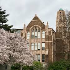 Exterior of a red and cream bricked university building with gothic architectural accents, a cherry blossom tree in the foreground.