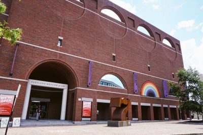 Exterior of a red bricked museum building, entrance sign reading "Portland Museum of Art." A Rainbow banner hangs between signs advertising exhibitions, and a large metal sculpture stands in front.