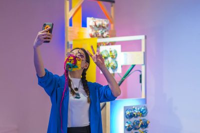 Person wearing glasses and a yellow face mask with sculptural hair extension attachments takes a selfie in front of a blurred art installation in the background.