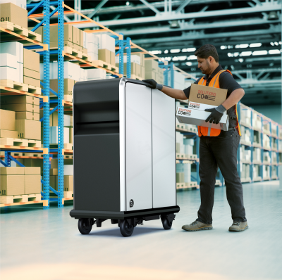 Dark skinned person wearing an orange safety vest in a warehouse surveys a wheeled black and white machine, holding a stack of boxes..
