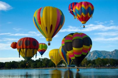 A group of 8 hot air balloons flying over a lake, a small mountain and forest in the background.
