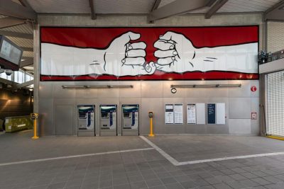 Interior of the entrance to a light rail station above ground. A public artwork mural of two white graphic hands pinky-swearing against a red background is above ticketing kiosks and information signs.