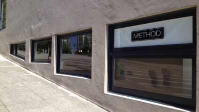 Exterior of four gallery windows at street left, part of the sidewalk visible. Sign on the outside of the closest window reads, "METHOD."
