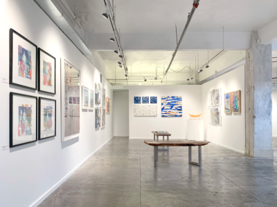 White walled rectangular gallery space showing several different types of artwork on the walls, as well as some three dimensional sculptures. Poured concrete floors.