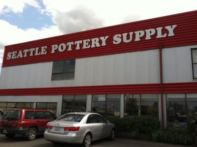 Exterior of a storefront, sign reading "Seattle Pottery Supply". The building is lined with windows, and two cars are parked in front.