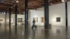 Interior of a white walled art gallery, featuring works of art on the walls in the background. A blurred, indistinct figure moves through the space.