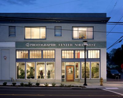 White brick building containing a photography gallery is lit up at night, several patrons visible through the storefront windows. Painted text on the building reads "Photographic Center Northwest."