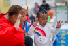 Dark skinned toddler wearing a rainbow sweatshirt makes a pretend scary face, smiling slightly, holding up outstretched fingers. A dark skinned adult wearing a bright red shirt to her left makes the same expression and gesture.
