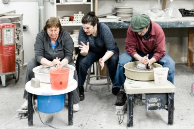 Two adults use pottery wheels to throw ceramic artwork while listening to a third adult standing between them, offering instruction.