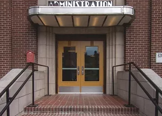Close up image of a yellow front door to a red brick building, red brick steps leading up to the door. A silver metal sign on the awning above the door reads "Administration"