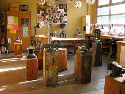 Sun shines through a large window of a small jewelry working studio. Several small anvils sit in the center of the studio space, with a table and cabinets in the back.