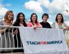 A group of five people stand in front of a white railing, holding a white banner that reads "Teach For America; www.teachforamerica.org".