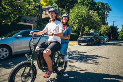 Two people wearing helmets ride an electric bicycle down the street, both smiling