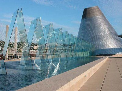 Glass art installation in a pool of water. In the background is a modern, geometric steel art museum building.