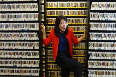 Femme person in a red sweater smiles widely while standing between shelves of CDs in a library setting.