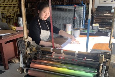 Femme person wearing an artist apron works at a letterpress