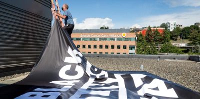 Facilities worker installs a large black banner on a building roof that reads "Black Lives Matter".